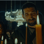Still from a film showing a man and women in a dark room with a chandelier behind them and candles in front. The man is smiling as he looks at the candles; the woman is looking across at him with a hint of a smile.