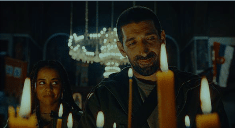 Still from a film showing a man and women in a dark room with a chandelier behind them and candles in front. The man is smiling as he looks at the candles; the woman is looking across at him with a hint of a smile.