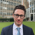 Image of a white man wearing glasses in a dark blue suit, white shirt and light blue tie with a background of tall office buildings and grass.